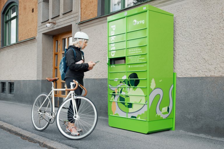 En kvinna står med en cykel framför en Bring paketbox och har öppnat en lucka med telefonen