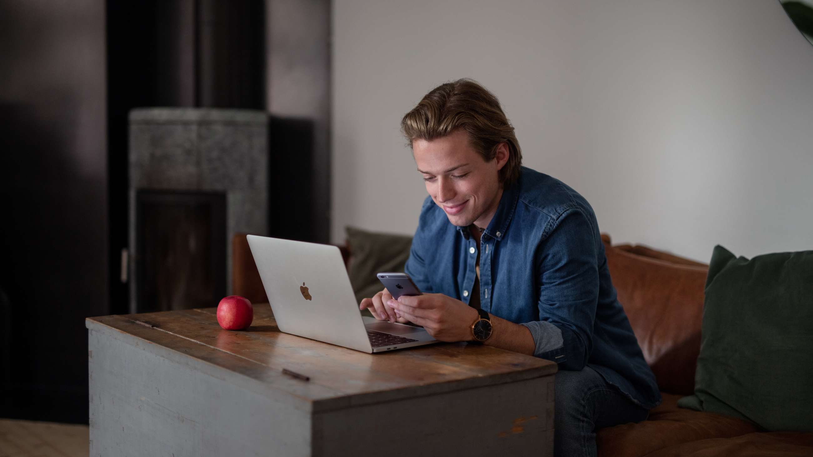 En man iklädd jeansskjorta sitter i en soffa framför en bärbar dator med en mobiltelefon i handen som han tittar på