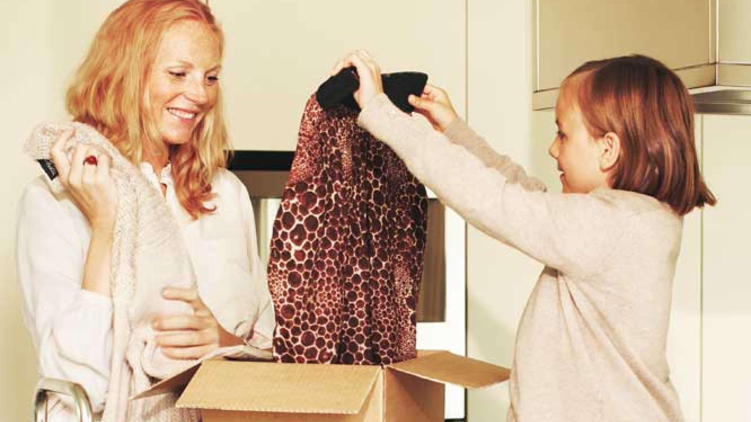En kvinna står och håller i en tröja framför ett barn som lyfter upp en annan tröja från ett paket som står mellan dem