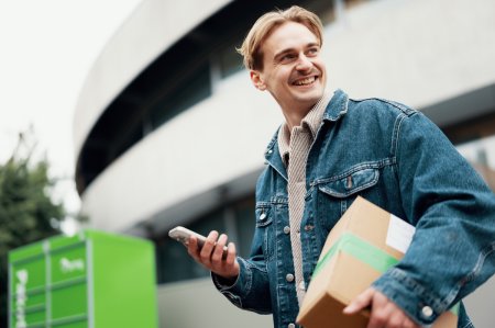 En man står vid en grön Bring-paketbox med en telefon och ett paket i händerna