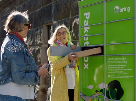 Två äldre damer står framför en grön Bring-paketbox och tar ett paket ur en lucka