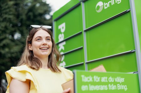 En leende kvinna med solglasögon på huvudet står framför en grön Bring-paketbox och hämtar ett paket från en öppen lucka
