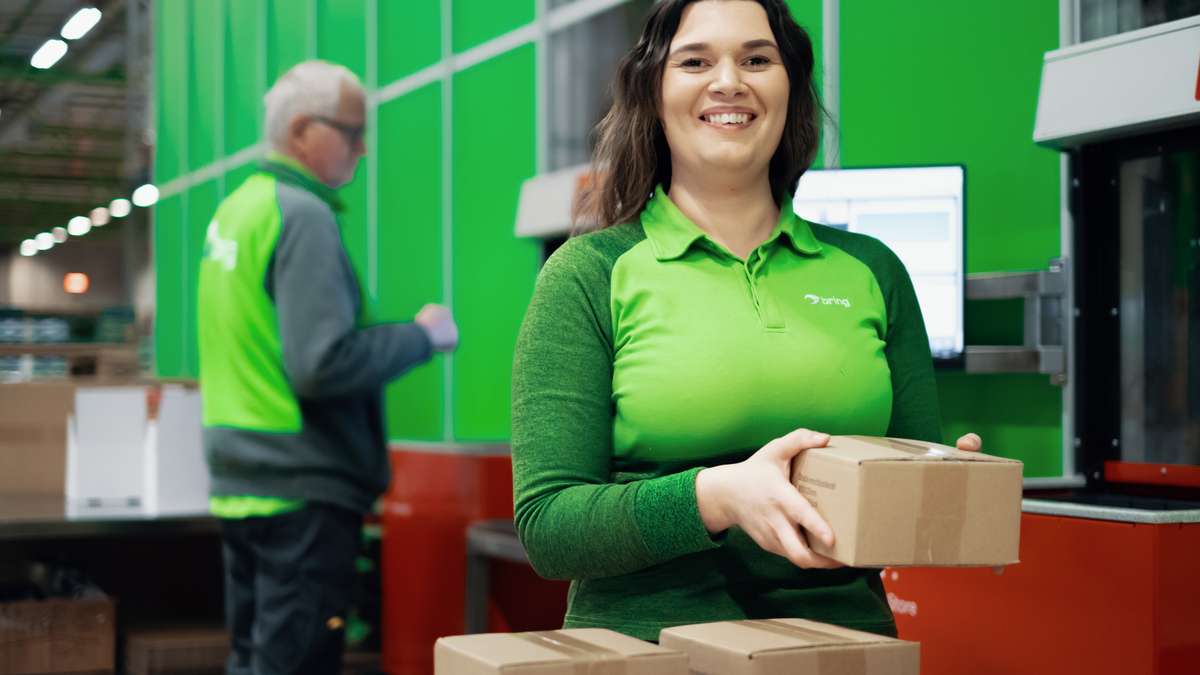 En kvinna står på ett lager och håller i ett paket samtidigt som hon ler mot kameran