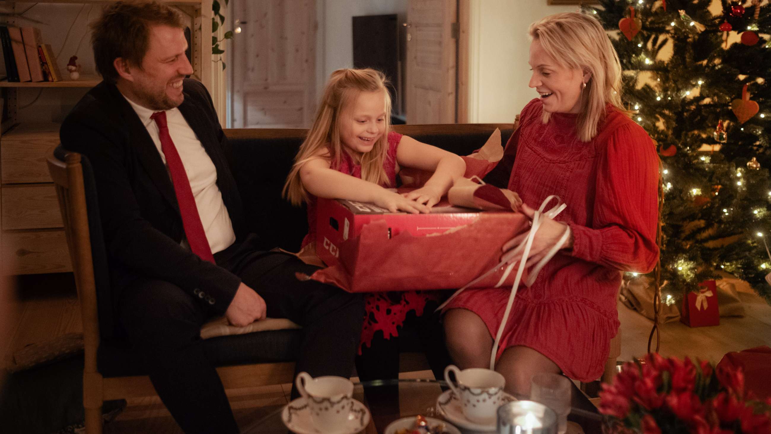 En barn sitter mellan en man och en kvinna på en soffa på julafton och öppnar en julklapp