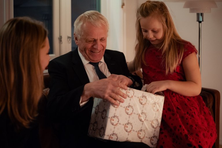 En äldre man sitter på en soffa med sitt barnbarn bredvid och öppnar en julklapp