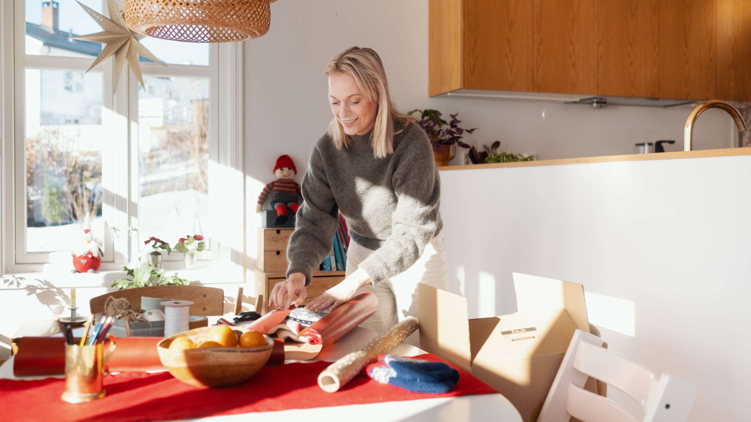 En kvinna står lutad över ett bord och ler och packar en tröja som en julklapp