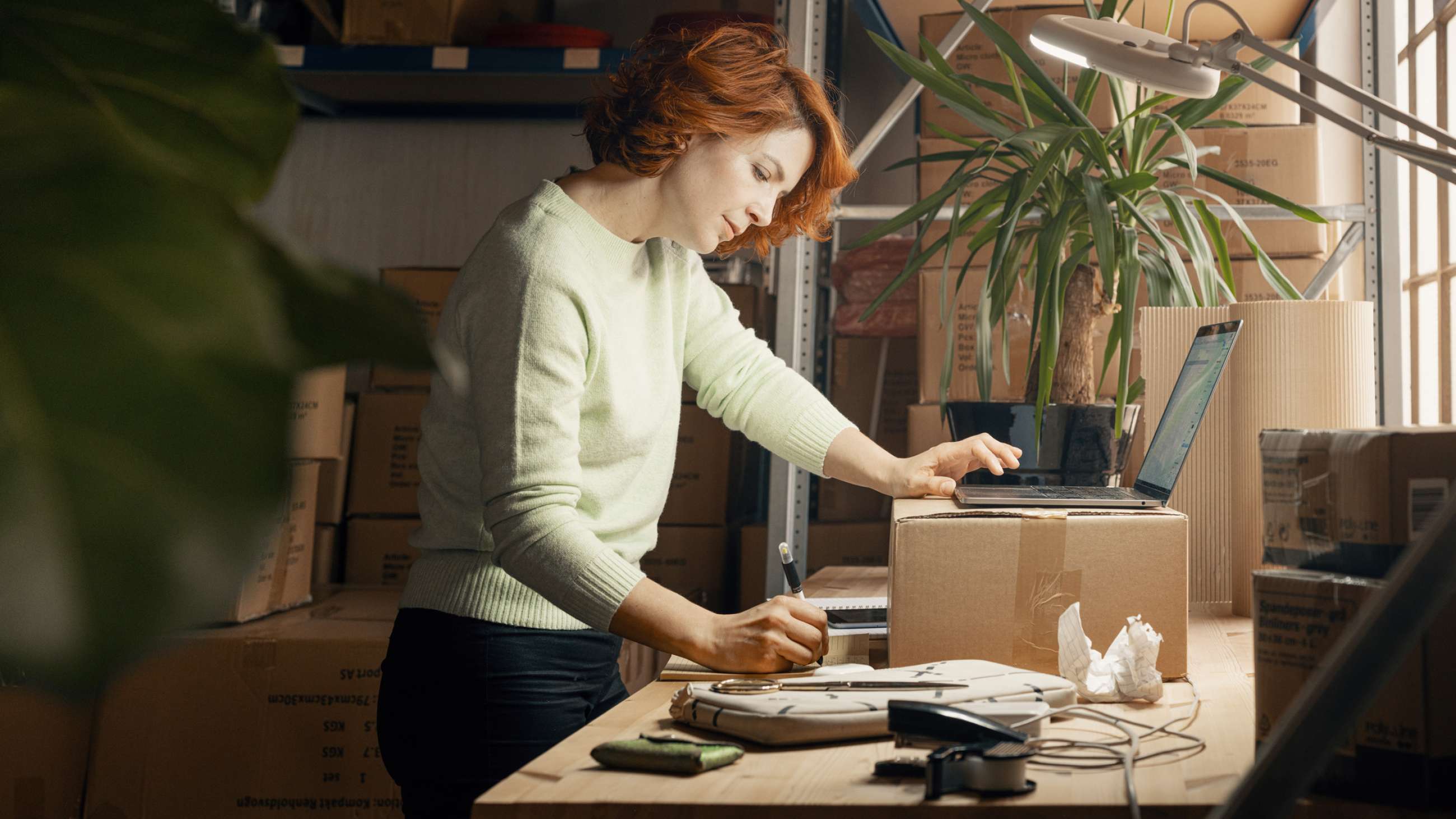 En kvinnlig nätbutiksinnehavare står vid en papplåda på ett arbetsbord med många lådor i bakgrunden. Hon antecknar i ett block medan hon håller den andra handen på en öppen laptop.