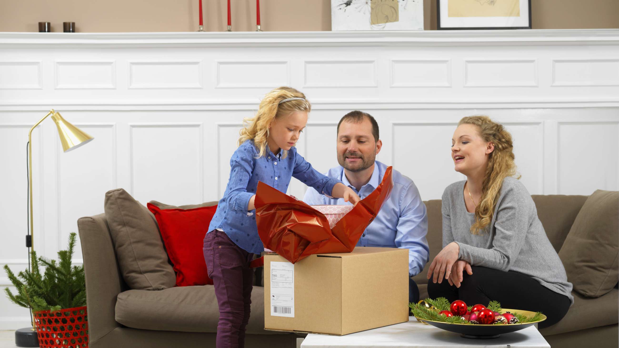 En man och en kvinna sitter på en soffa medan en liten tjej öppnar ett paket på bordet som är inslagen som en julklapp