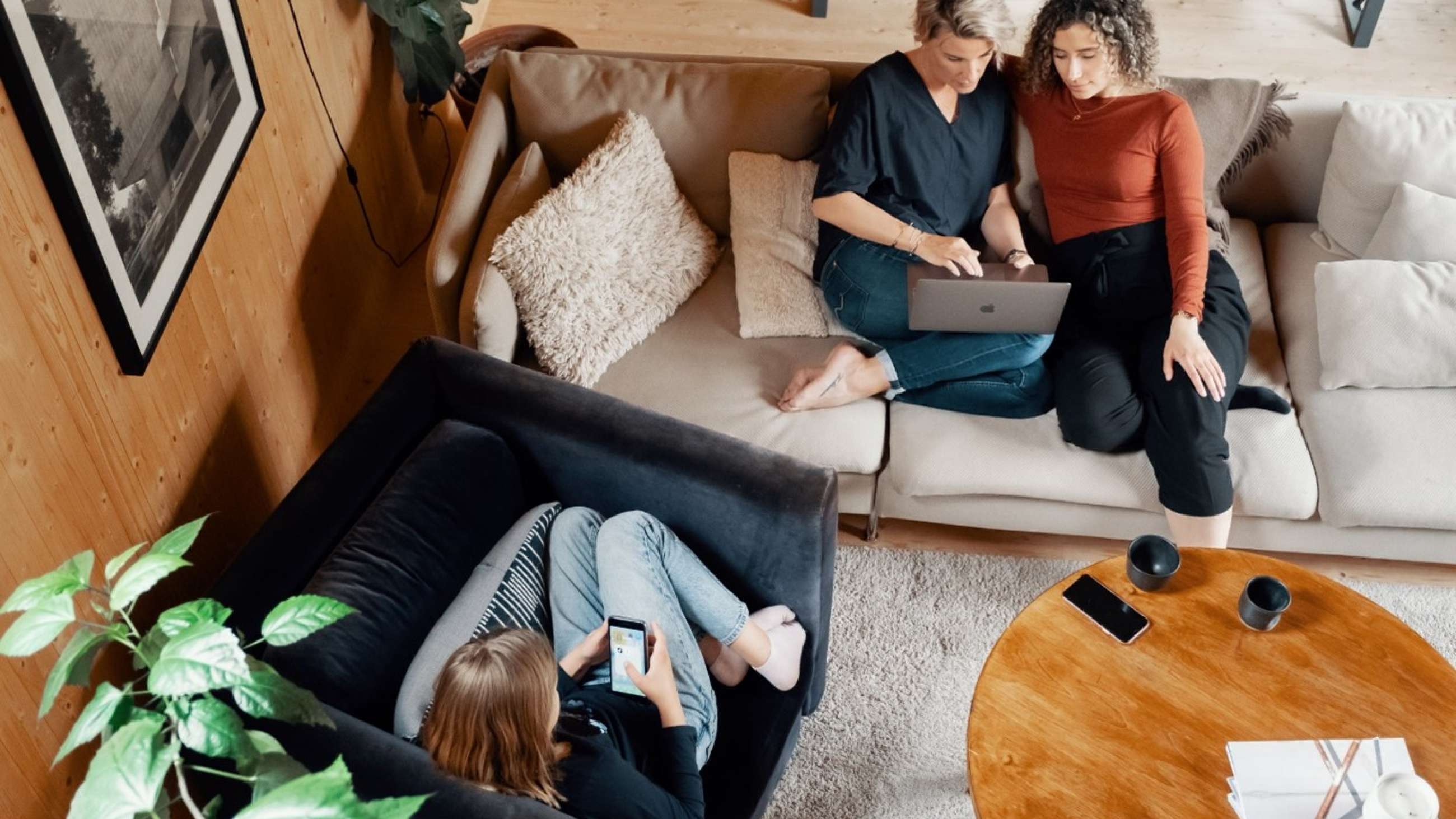 Tre personer i ett vardagsrum, sittandes med mobiltelefon och laptop
