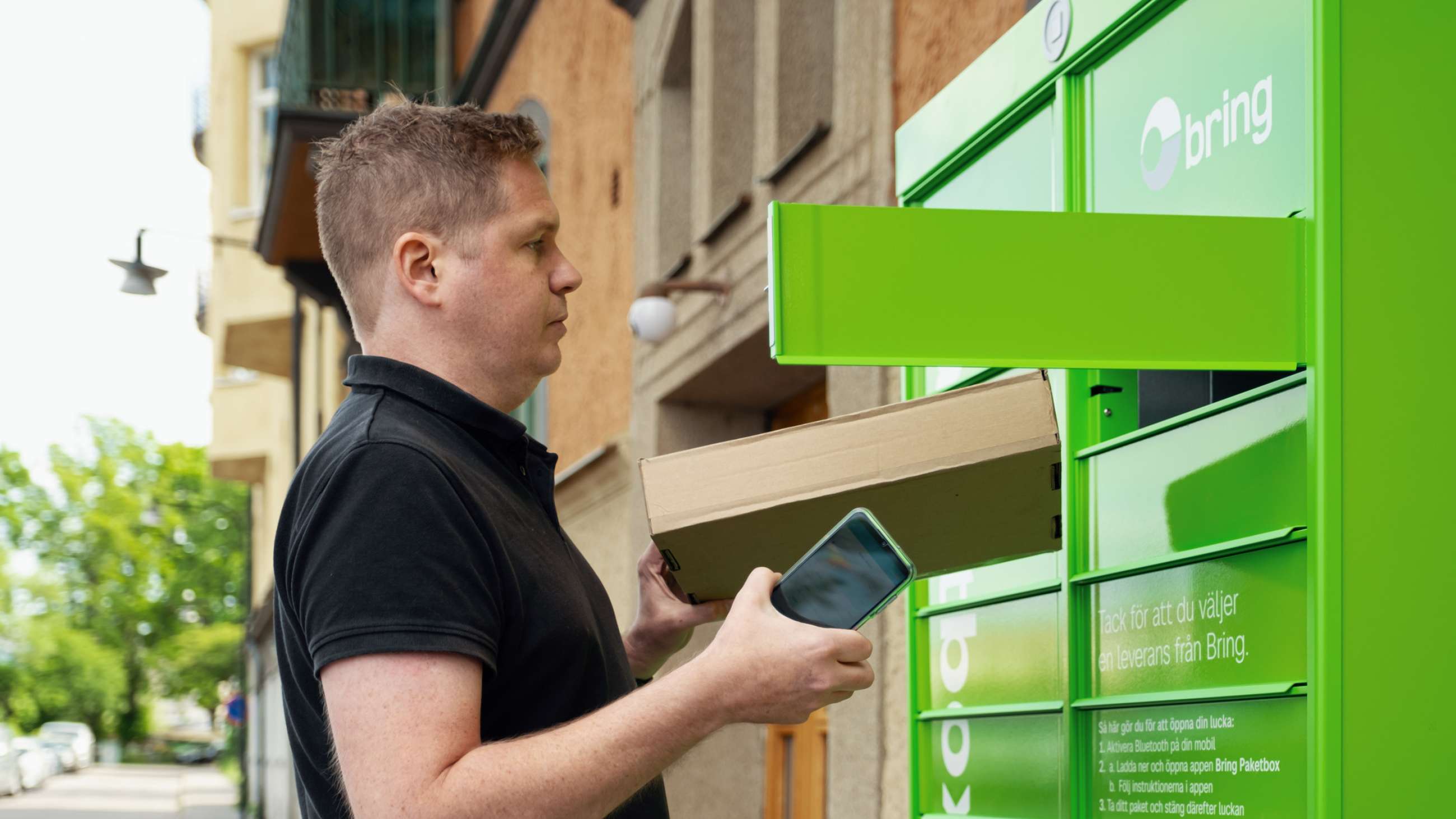 En man står med en mobil i handen framför en grön Bring paketbox och lämnar ett paket för retur i en lucka i boxen