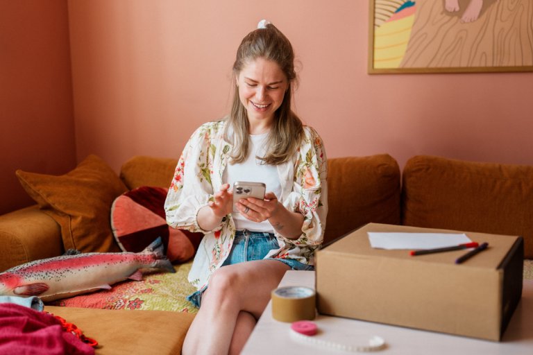 Tjej sitter i soffan med ett paket bredvid sig och håller i sin mobil
