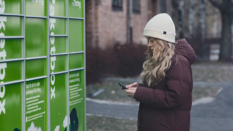 En tjej står vid en Bring paketbox med mobilen i handen och ska hämta ut ett paket