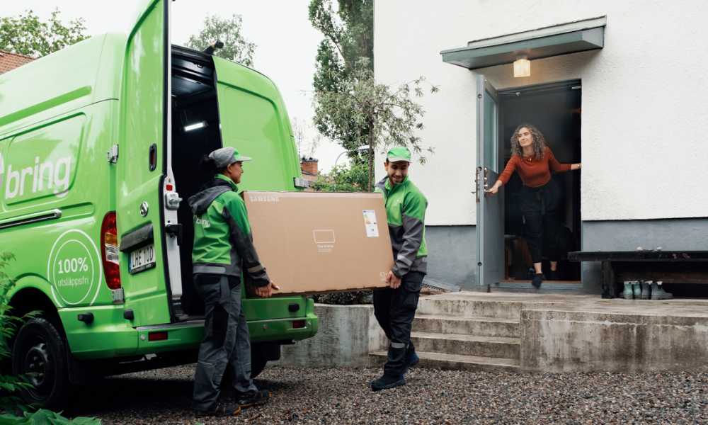 Två Bring chaufförer bär in ett stort paket till en kvinna som öppnar en dörr till ett hus.
